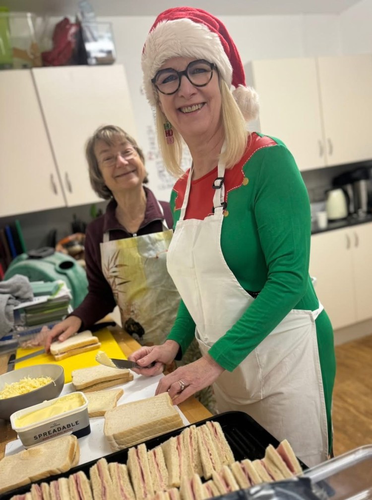 Kay making sandwiches for the Hub party. Shows ham sandwiches in a row and Kay with bread and a knife, wearing a Santa hat. Lesley, another helper, looks on. 