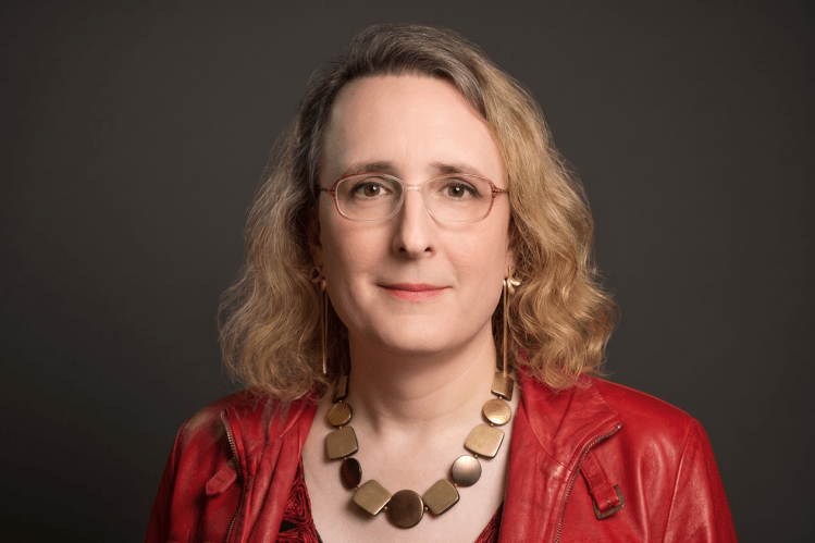 A headshot of Abby looking straight at the camera. She has blond hair, wears glasses, and is wearing a read jacket.