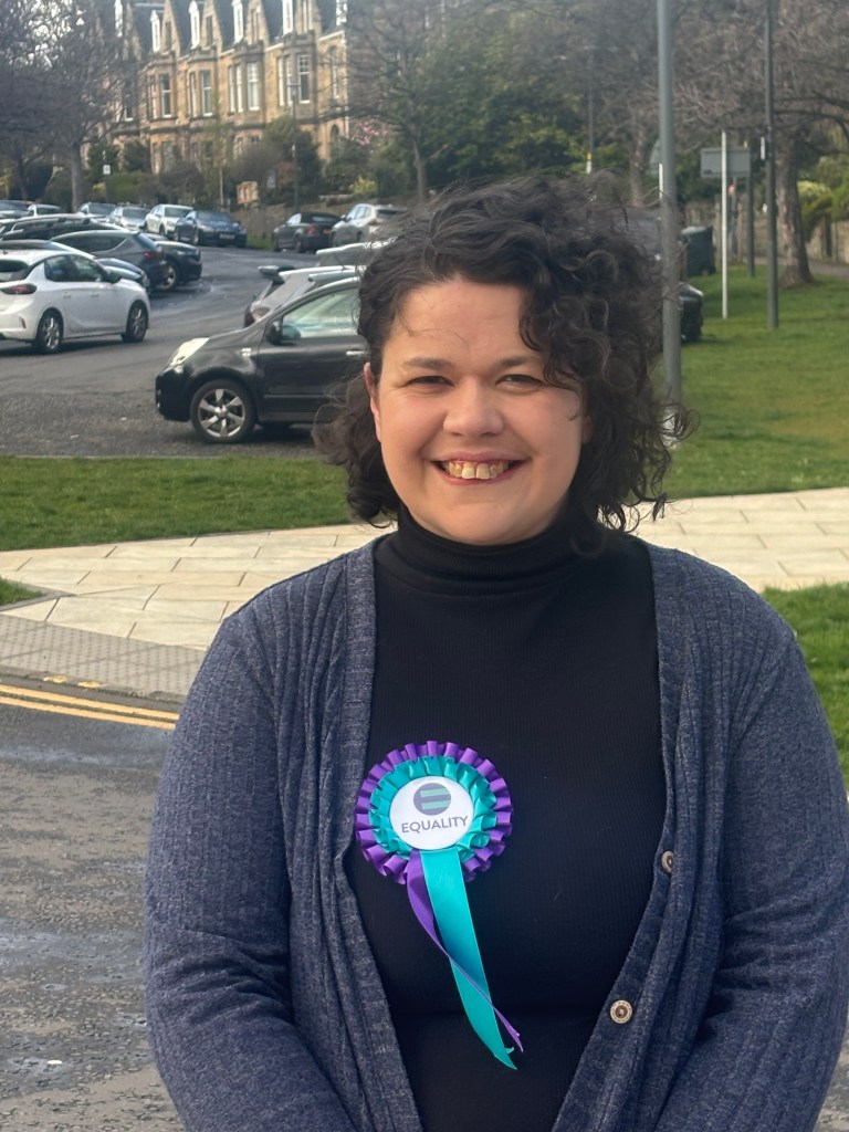 Photo of the dark-haired Laura Mackintosh wearing an Equality Party rosette.