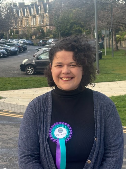 Picture of Laura, a woman in her 30s with dark hair and an Equality Party rosette, standing on an Edinburgh street.
