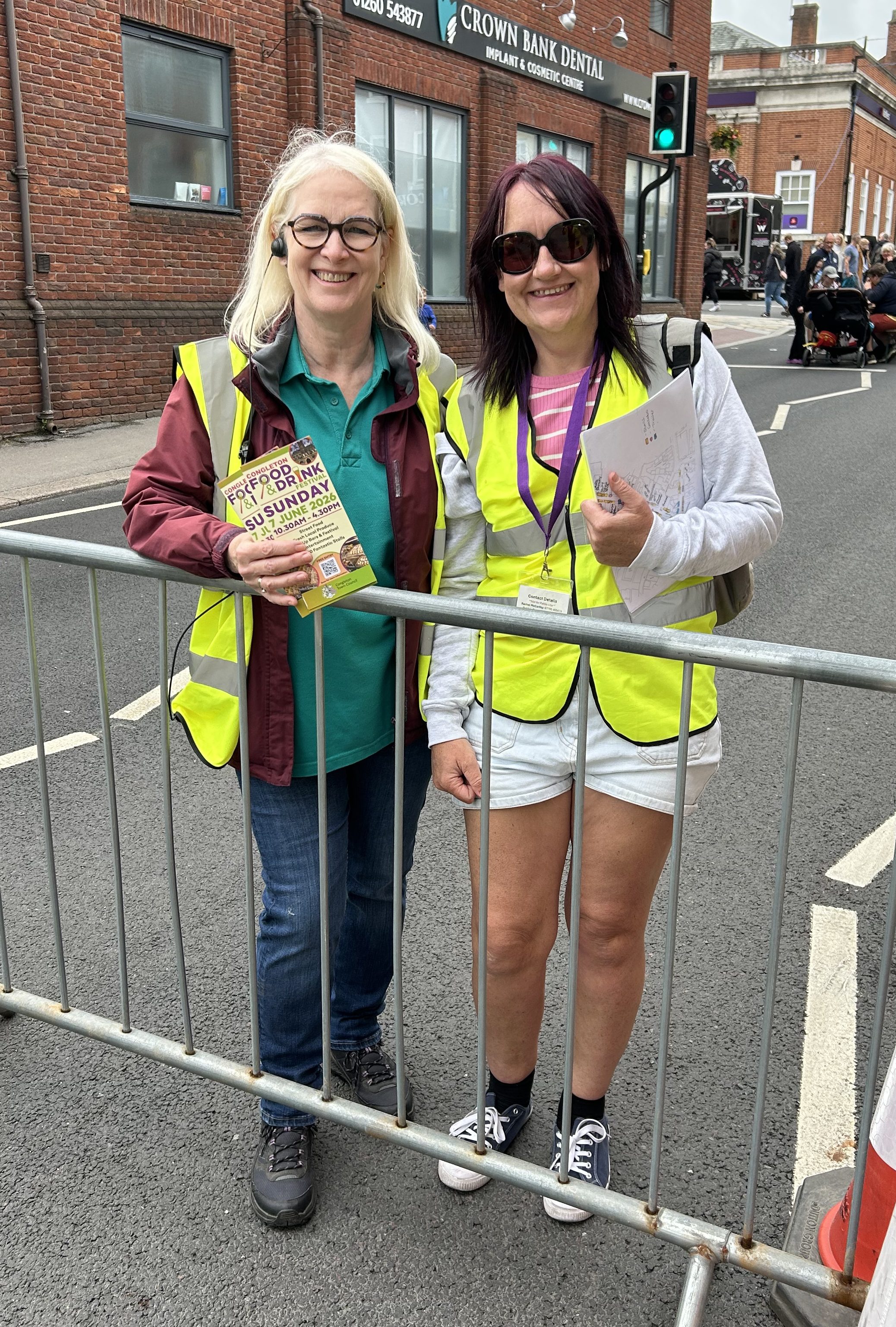 Blond Kay and dark-haired Susie in hi viz jackets at a road barrier.