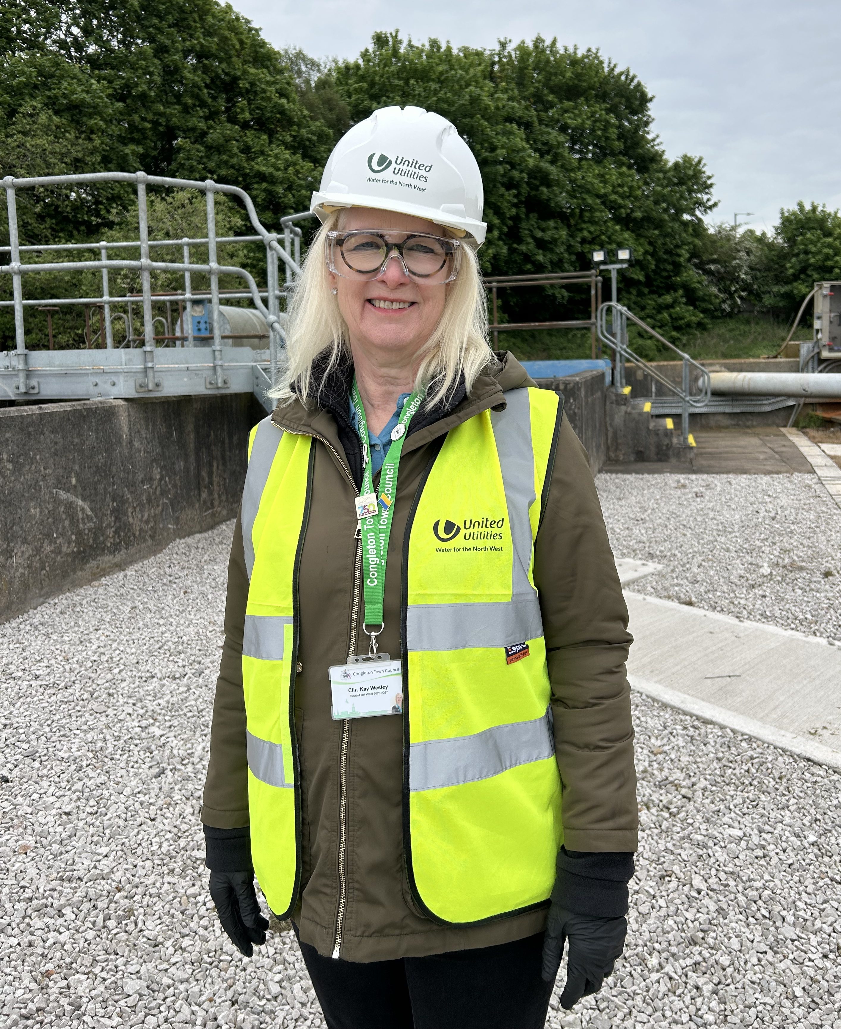 Kay with hi viz, safety glasses and hard hat, with industrial-looking fencing behind.
